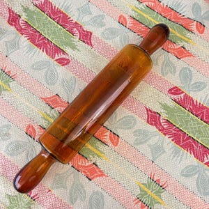 May include: Amber-colored glass rolling pin with rounded handles. The rolling pin is positioned diagonally on a patterned fabric with stripes of pink, green, and white floral designs. A kitchen utensil for baking.
