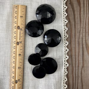 May include: A collection of seven black, faceted and smooth, round buttons of varying sizes. A wooden ruler with inch measurements is placed next to the buttons. The buttons are on a white fabric with a lace trim.