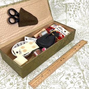 Op de afbeelding: Een open olijfgroene naaidoos gevuld met naaispullen. De doos bevat een zwarte schaar, klosjes garen, knopen, een kleine zwarte zak en een houten liniaal. De liniaal is gemarkeerd met centimeters.