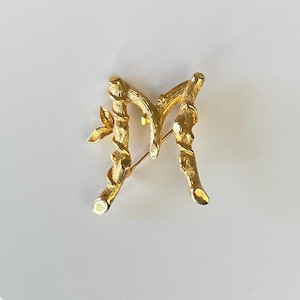 May include: A gold-toned metal brooch in the shape of the letter "M". The brooch is designed to resemble intertwined branches with small leaves. The pin is secured with a gold-colored clasp.