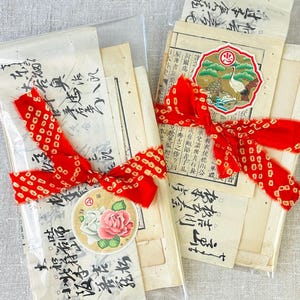 May include: Two clear plastic sleeves containing stacks of vintage paper with black Japanese calligraphy. Each bundle is tied with a bright red ribbon featuring a white dot pattern. One bundle has a round rose sticker, the other a crane sticker.