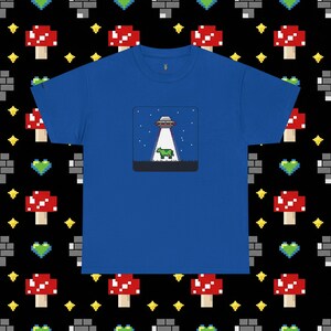 May include: A royal blue t-shirt featuring a pixelated graphic of a UFO beaming a green cow. The design is set within a square frame. The background is a repeating pattern of pixelated mushrooms, bricks, and stars.