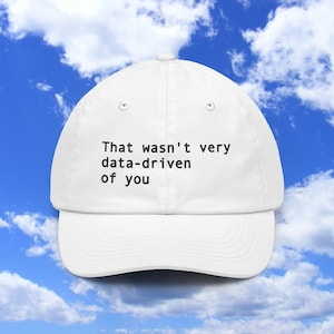 May include: A white baseball cap featuring the black text "That wasn't very data-driven of you" printed on the front. The cap has a structured crown, a curved brim, and a button at the top center, presented against a bright blue sky background.