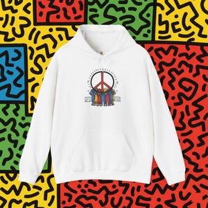 Op de afbeelding: Witte hoodie met een voorzak. De afbeelding op de voorkant toont een vredesteken met een rode kern, omringd door een wijzerplaat en figuren in verschillende kleuren. De achtergrond heeft een kleurrijk, abstract patroon.