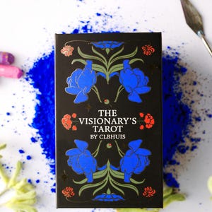 May include: A black tarot card box with blue floral illustrations and the text "THE VISIONARY'S TAROT BY CLBHUIS" is surrounded by blue pigment, paint tubes, and art supplies. The box features gold accents and red floral details.