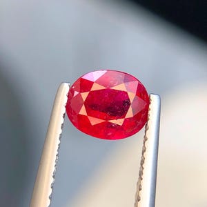 K&ouml;nnte beinhalten: Ein oval geschliffener, leuchtend roter Edelstein, der von einer silberfarbenen Metallpinzette gehalten wird. Der Stein hat eine facettierte Oberfl&auml;che, die das Licht reflektiert und einen tiefen Karmesinton sowie eine klare Transparenz zeigt.