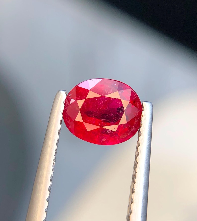 K&ouml;nnte beinhalten: Ein oval geschliffener, leuchtend roter Edelstein, der von einer silberfarbenen Metallpinzette gehalten wird. Der Stein hat eine facettierte Oberfl&auml;che, die das Licht reflektiert und einen tiefen Karmesinton sowie eine klare Transparenz zeigt.