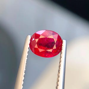 Puede incluir: Una gema roja de corte ovalado sostenida por pinzas de metal plateado. La piedra presenta una superficie facetada que refleja la luz, mostrando un tono carmesí profundo y saturado en todo su cuerpo translúcido y una apariencia pulida.