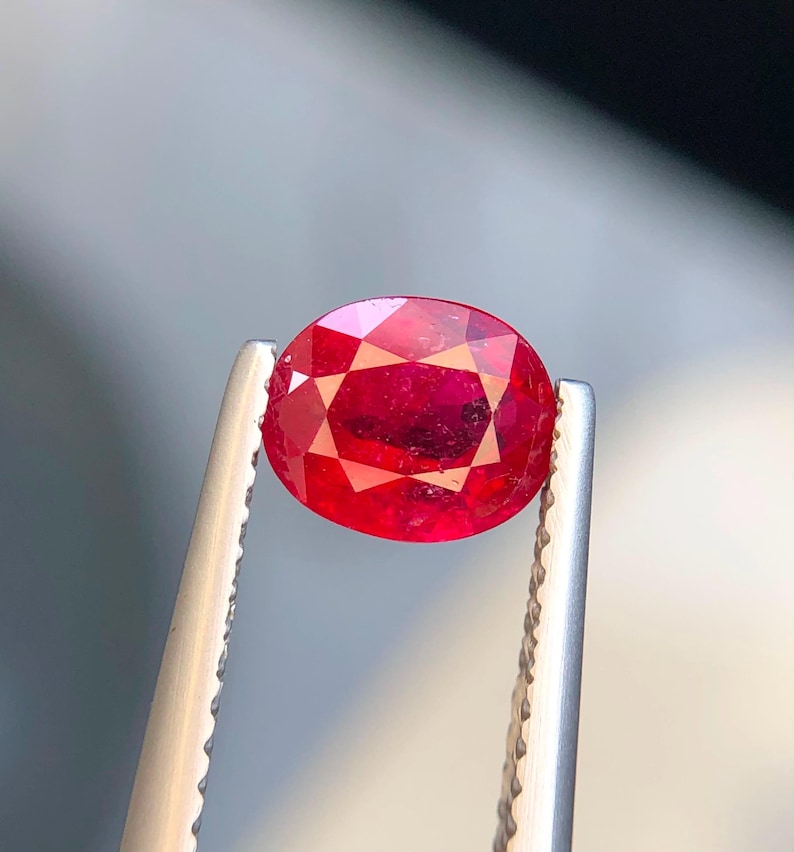 K&ouml;nnte beinhalten: Ein leuchtend roter Edelstein im Ovalschliff, gehalten von einer silberfarbenen Metallpinzette. Der Stein besitzt eine facettierte Oberfl&auml;che, die das Licht reflektiert und einen tiefen, ges&auml;ttigten Karmesinton im gesamten durchscheinenden K&ouml;rper zeigt.