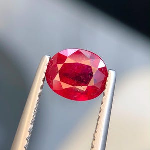 K&ouml;nnte beinhalten: Ein leuchtender, oval geschliffener roter Edelstein, der von einer silberfarbenen Metallpinzette gehalten wird. Der Stein hat eine facettierte Oberfl&auml;che, die das Licht reflektiert und einen tiefen, satten Karmesinton zeigt.