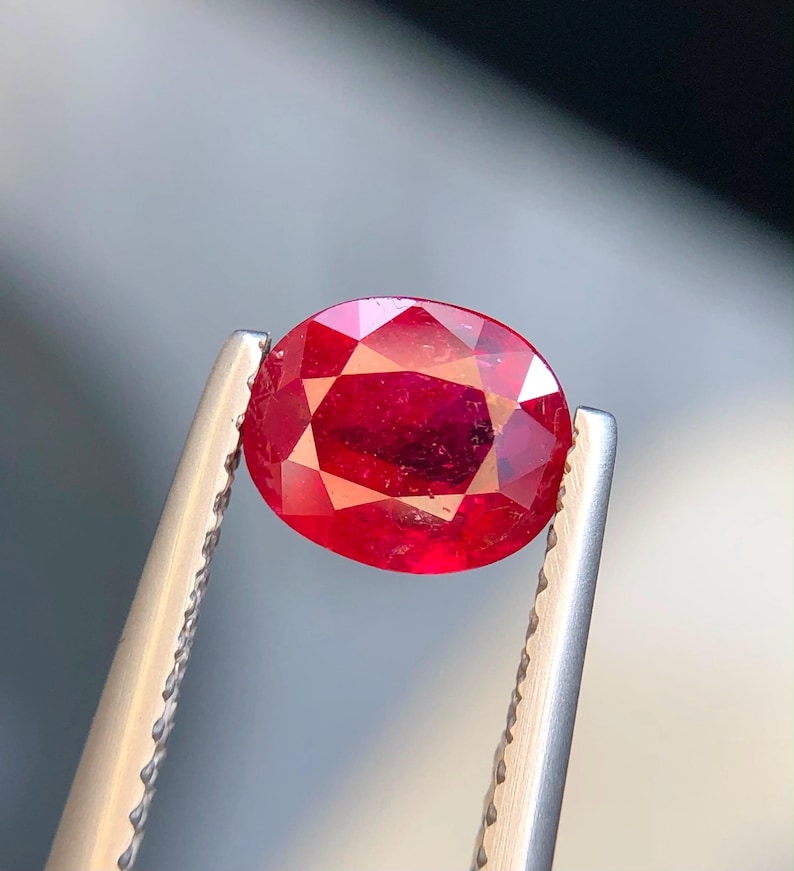 K&ouml;nnte beinhalten: Ein leuchtender, oval geschliffener roter Edelstein, der von einer silberfarbenen Metallpinzette gehalten wird. Der Stein hat eine facettierte Oberfl&auml;che, die das Licht reflektiert und einen tiefen, satten Karmesinton zeigt.