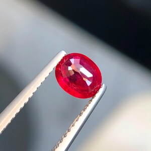 K&ouml;nnte beinhalten: Ein leuchtend roter Edelstein im Ovalschliff, gehalten von einer silberfarbenen Metallpinzette. Der Stein besitzt eine facettierte Oberfl&auml;che, die das Licht reflektiert und einen tiefen, ges&auml;ttigten Karmesinton im transparenten K&ouml;rper zeigt.