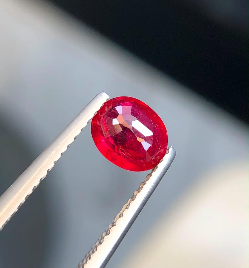 K&ouml;nnte beinhalten: Ein leuchtend roter Edelstein im Ovalschliff, gehalten von einer silberfarbenen Metallpinzette. Der Stein besitzt eine facettierte Oberfl&auml;che, die das Licht reflektiert und einen tiefen, ges&auml;ttigten Karmesinton im transparenten K&ouml;rper zeigt.