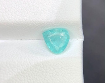 Könnte beinhalten: Ein loser, facettierter Edelstein in dreieckiger Form mit einer leuchtenden, durchscheinenden cyanblauen Farbe. Der Stein weist mehrere geometrische Facetten auf, die das Licht reflektieren und auf einer strukturierten weißen Oberfläche funkeln.