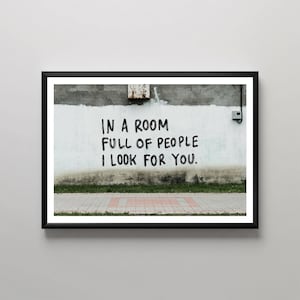 Op de afbeelding: Ingelijst kunstwerk met de tekst "IN A ROOM FULL OF PEOPLE I LOOK FOR YOU" in zwart op een witte muur. De muur heeft een verweerde uitstraling, met gras en een rode bakstenen stoep eronder. De lijst is zwart.