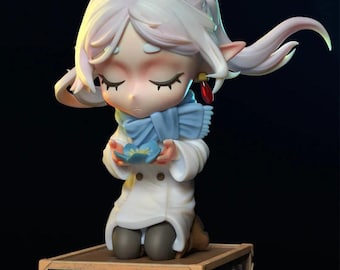 May include: A figurine of a kneeling character with closed eyes, wearing a white coat and blue scarf, holding a blue flower. The figure has long white hair and is kneeling on a wooden box with blue flowers around it.