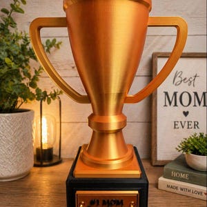 May include: A gold-colored trophy with a black base. The trophy has two handles and a plaque that reads "#1 MOM From Brettie". A sign in the background says "Best MOM Ever".