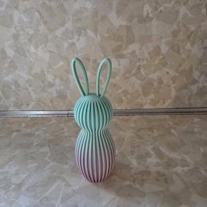 May include: A decorative bunny figurine with a ribbed design. The top portion is a light teal color, transitioning to a pinkish-purple at the base. The bunny has long, curved ears.