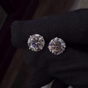 May include: Two round-cut diamond stud earrings. The diamonds are clear and sparkling, set against a dark background. The earrings are shown close up, highlighting their brilliance and clarity. The earrings are a classic jewelry item.