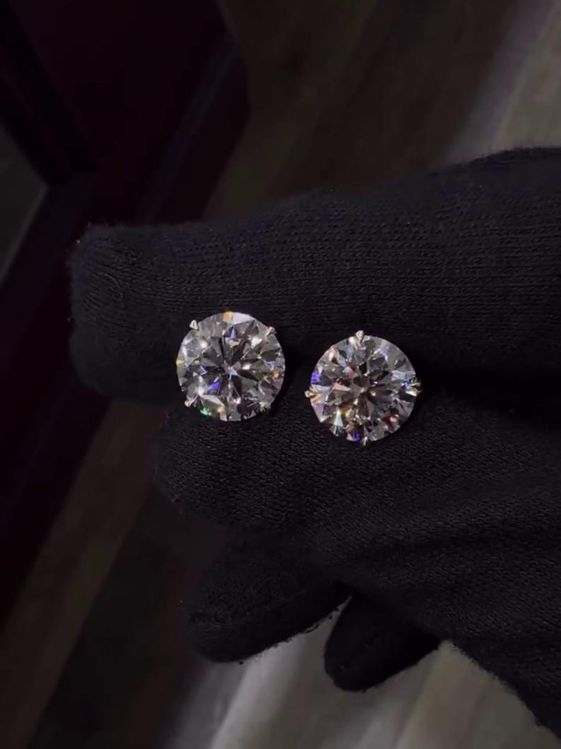 May include: Two round-cut diamond stud earrings. The diamonds are clear and sparkling, set against a dark background. The earrings are shown close up, highlighting their brilliance and clarity. The earrings are a classic jewelry item.