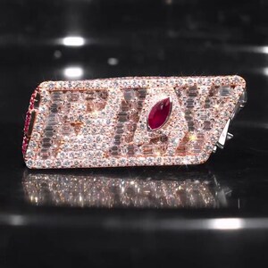 Può includere: Un anello rettangolare con la scritta "SAV" in piccole pietre trasparenti. L'anello è incrostato di piccole pietre trasparenti e presenta una grande gemma rossa di forma ovale al centro. L'anello è appoggiato su una superficie nera.