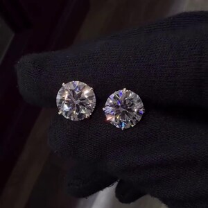 May include: Two round, brilliant-cut diamond stud earrings. The diamonds are clear and sparkle, set against a dark background. The earrings are held by a gloved hand, showcasing their size and brilliance. A classic jewelry item.