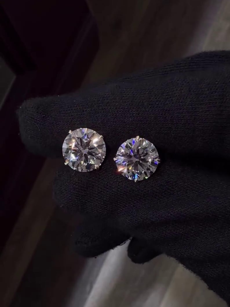 May include: Two round, brilliant-cut diamond stud earrings. The diamonds are clear and sparkle, set against a dark background. The earrings are held by a gloved hand, showcasing their size and brilliance. A classic jewelry item.