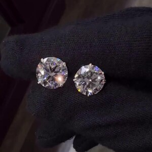 May include: Two round-cut diamond stud earrings. The diamonds are set in a silver-colored metal and are displayed against a black background. The earrings are sparkling and reflect light.
