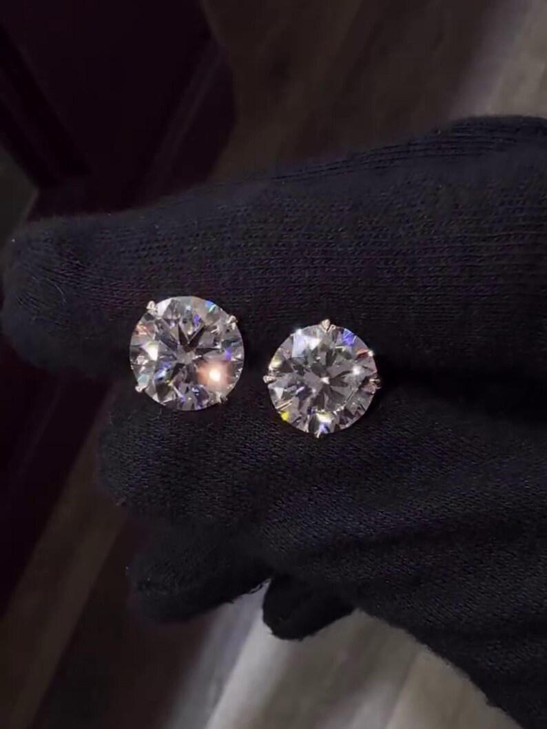 May include: Two round-cut diamond stud earrings. The diamonds are set in a silver-colored metal and are displayed against a black background. The earrings are sparkling and reflect light.