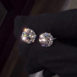 May include: Two sparkling, round-cut diamond stud earrings. The diamonds are held in a six-prong setting, reflecting light and creating a brilliant display. The earrings are set against a dark background.