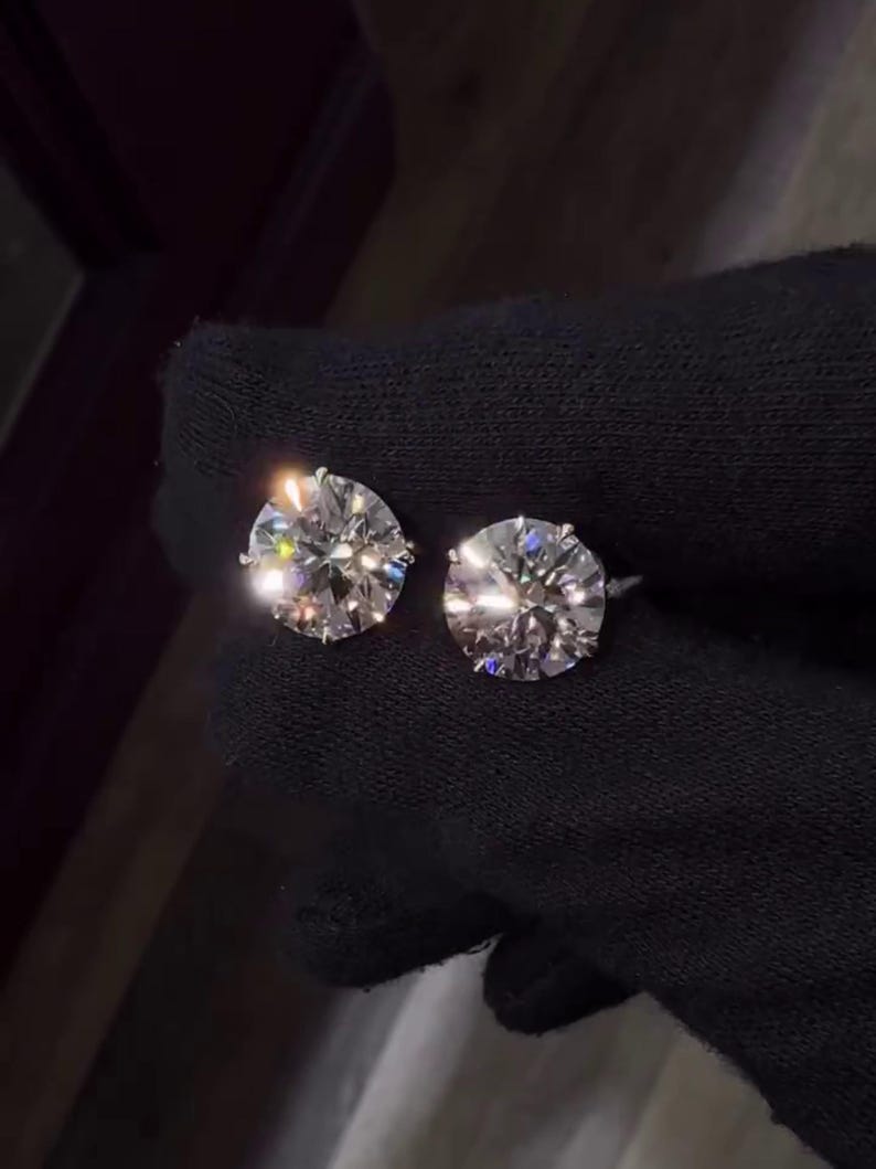 May include: Two sparkling, round-cut diamond stud earrings. The diamonds are held in a six-prong setting, reflecting light and creating a brilliant display. The earrings are set against a dark background.