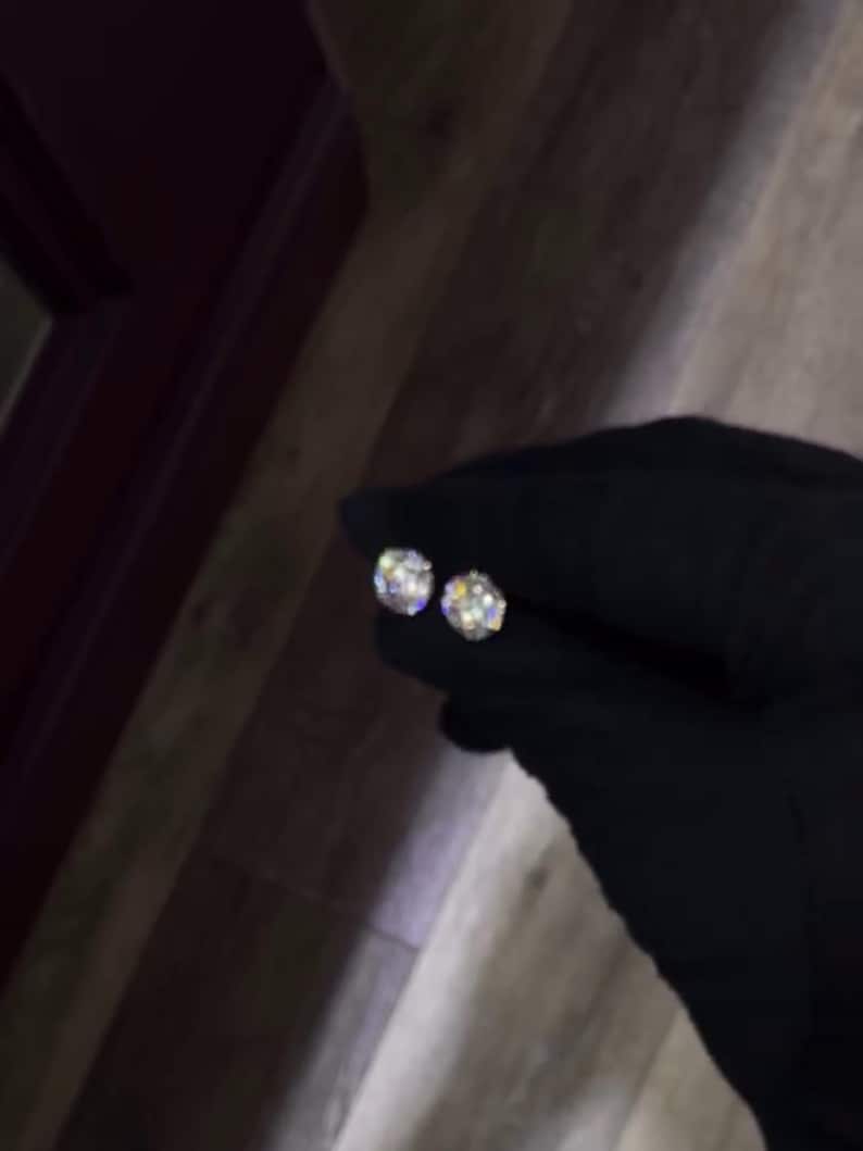 May include: Two round, sparkling diamond stud earrings. The earrings are held by a gloved hand, showcasing their brilliance. The diamonds reflect light, creating a dazzling display. A classic jewelry item.