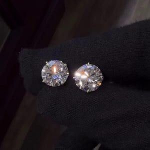 May include: Two round-cut diamond stud earrings. The diamonds are clear and sparkling, set against a dark background. The earrings are shown in a close-up view, highlighting their brilliance and clarity. The setting appears to be a classic prong style.