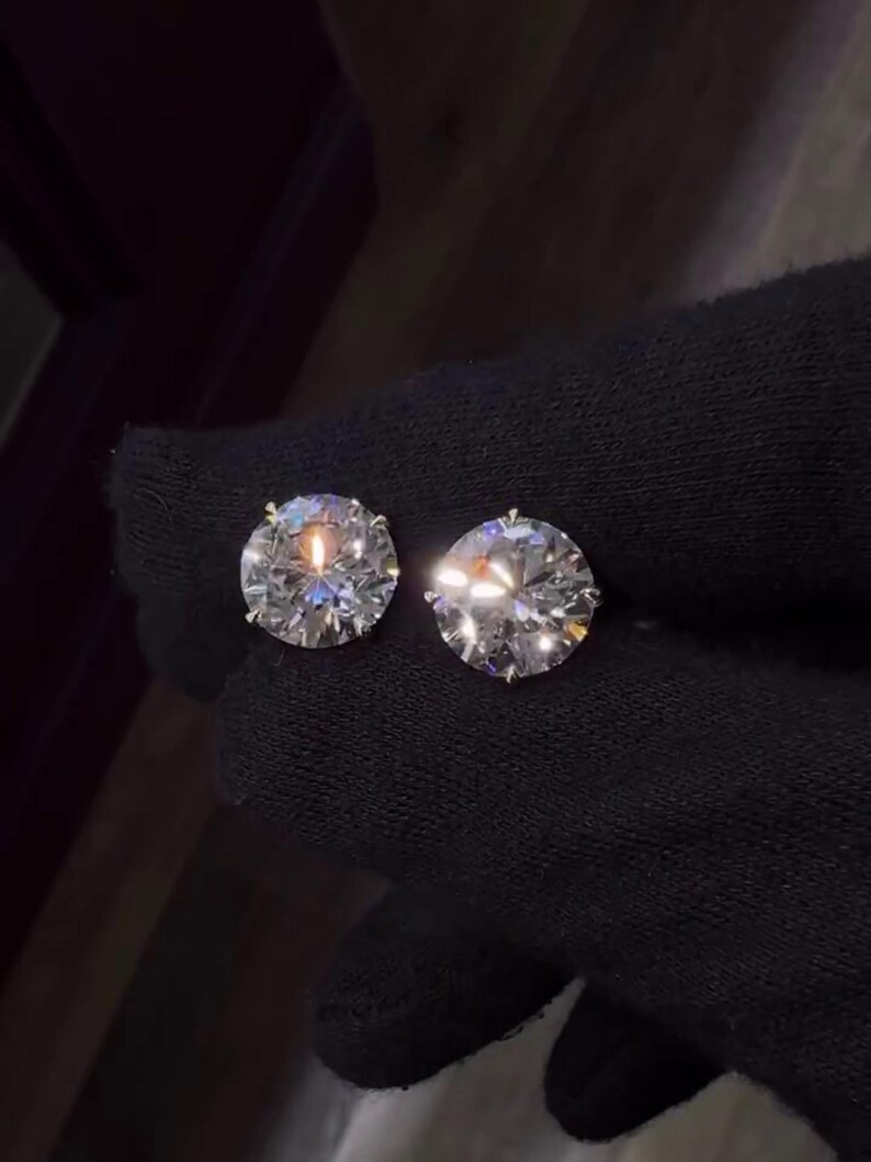 May include: Two round-cut diamond stud earrings. The diamonds are clear and sparkling, set against a dark background. The earrings are shown in a close-up view, highlighting their brilliance and clarity. The setting appears to be a classic prong style.