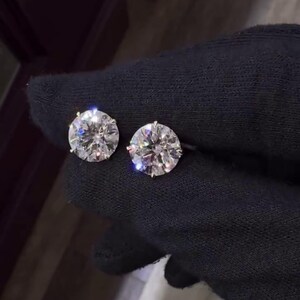 May include: Two round, brilliant-cut diamond stud earrings. The diamonds are set in a gold or gold-toned setting and are displayed against a black background. The earrings are shown close up, highlighting their sparkle and clarity.