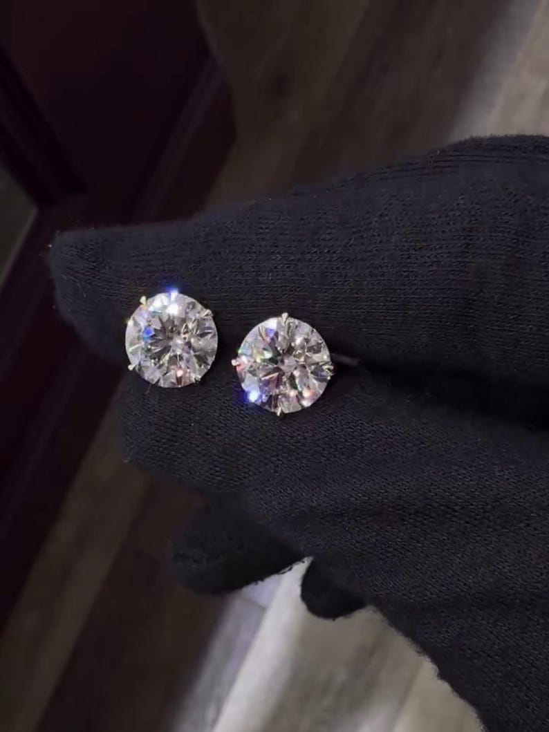 May include: Two round, brilliant-cut diamond stud earrings. The diamonds are set in a gold or gold-toned setting and are displayed against a black background. The earrings are shown close up, highlighting their sparkle and clarity.