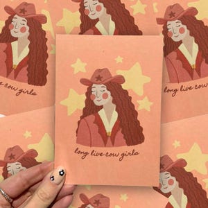 May include: Peach-coloured cards with a cowgirl illustration. The cowgirl has long, wavy red hair, a red cowboy hat, and a red jacket. The text "long live cow girls" is written on the card. Yellow stars are in the background.