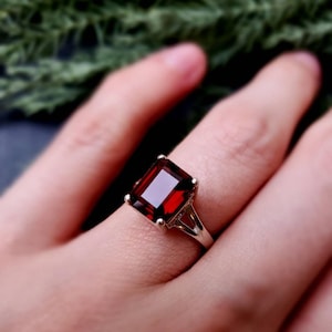 May include: A gold ring featuring a large, rectangular, deep red gemstone. The ring's band is a simple, elegant design, and the gemstone is set with four prongs. The ring is shown on a finger.