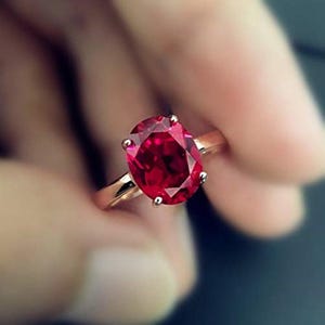 Może przedstawiać: Złoty pierścionek z dużym, owalnym, ciemnoczerwonym kamieniem szlachetnym. Kamień jest trzymany przez cztery pazury. Obręcz pierścionka ma ciepły złoty kolor, a kamień ma wiele faset, które odbijają światło.