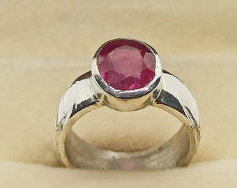 May include: A silver ring with a large, oval-cut ruby gemstone. The ruby is a deep red color and is set in a polished silver band. The ring is displayed in a beige jewelry box.