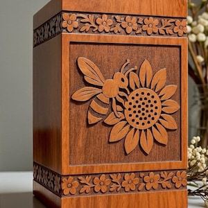 Può includere: Una scatola di legno con un disegno intagliato di ape e girasole. La scatola è di colore marrone caldo e presenta intagli floreali sulla parte superiore e inferiore. La scatola è appoggiata su una superficie bianca.