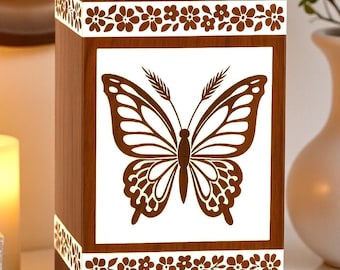 Puede incluir: Una urna rectangular de madera con un acabado de veta de madera marrón medio. El panel frontal presenta un gráfico grande de una mariposa marrón centrada sobre un fondo blanco, enmarcado por bandas blancas horizontales con un patrón floral.