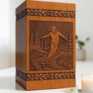 May include: A rectangular wooden cremation urn featuring a carved relief panel on the front. The carving depicts a person riding a skateboard on a winding path through a forest of pine trees. The top and bottom edges have a decorative leaf border.