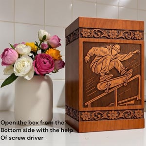 Wooden memorial urn box for ashes with skateboard engraved artwork, floral carved borders, polished natural wood texture, standing on shelf near candle and flowers, ideal for remembrance and tribute.