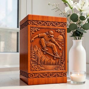 May include: A rectangular wooden urn featuring a detailed relief carving of a person skateboarding over a landscape with mountains, pine trees, and a setting sun. The top and bottom borders are decorated with intricate scrolling patterns.