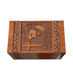 Elegant handcrafted wooden urn box engraved with the image of Jesus and a cross, featuring the words “Be Still and Know.” Made from solid wood with detailed carved floral borders and smooth finish. A beautiful Christian memorial urn for adult ashes, symbolizing peace, love, and eternal faith. Perfect for home remembrance or funeral service display.