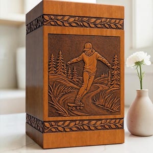 May include: A rectangular wooden urn with a medium brown finish. The front panel features a carved relief of a person skateboarding on a path through a forest of pine trees. The top and bottom borders are decorated with a carved leaf pattern.
