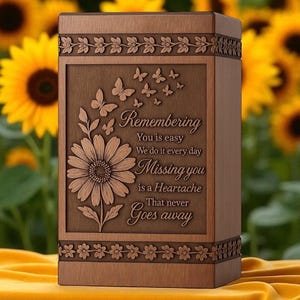 Puede incluir: Una urna de madera con un diseño floral y de mariposas. La urna tiene las palabras "Remembering", "Missing you is a Heartache That never Goes away" grabadas en la parte delantera. Girasoles en el fondo.