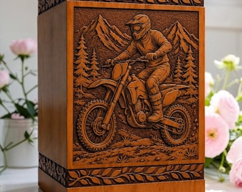 Könnte beinhalten: Eine rechteckige Urne aus Holz mit einer detaillierten Reliefschnitzerei einer Person auf einem Dirtbike in einer bergigen Waldlandschaft. Die oberen und unteren Kanten der Urne sind mit einem geschnitzten Blattmuster verziert.