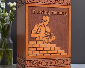May include: A rectangular wooden cremation urn featuring a carved relief of a bricklayer wearing a hard hat and overalls, working on a brick wall. The text "In loving memory" is engraved above the figure, framed by ornate scrollwork borders.